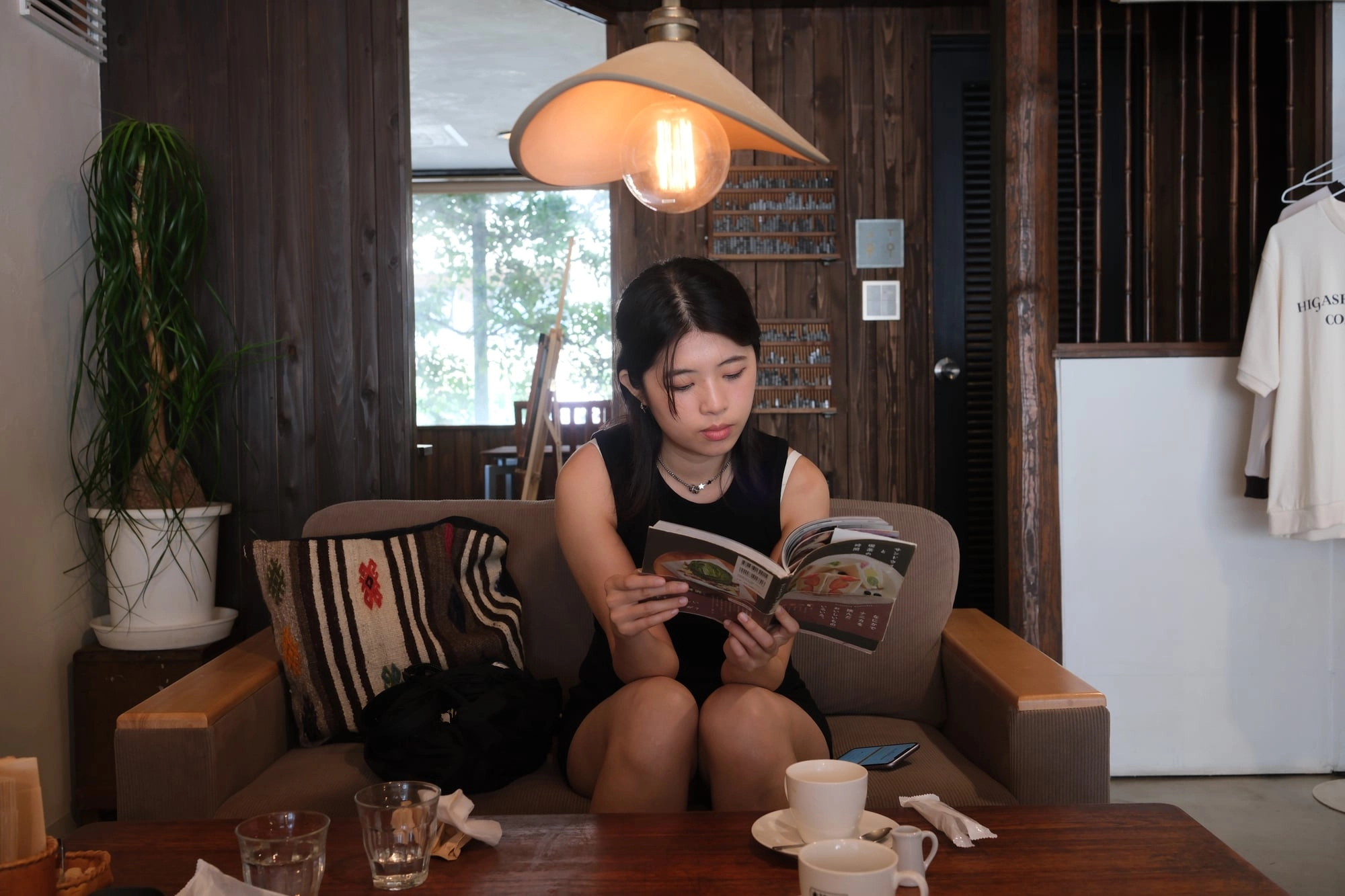 A young woman reads a recipe book on a sofa in a warm, wood-paneled café corner.