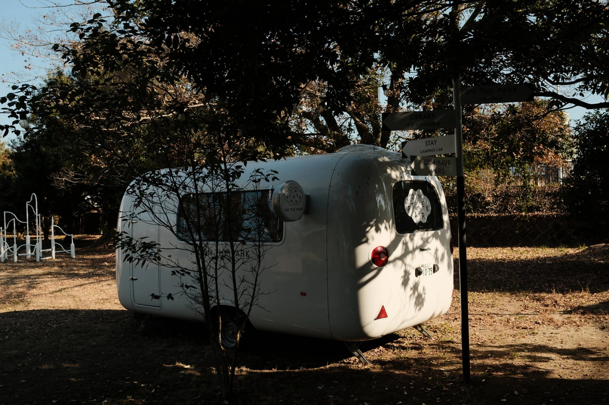 A vintage caravan trailer rests beneath bare trees, with signposts pointing toward nearby attractions.