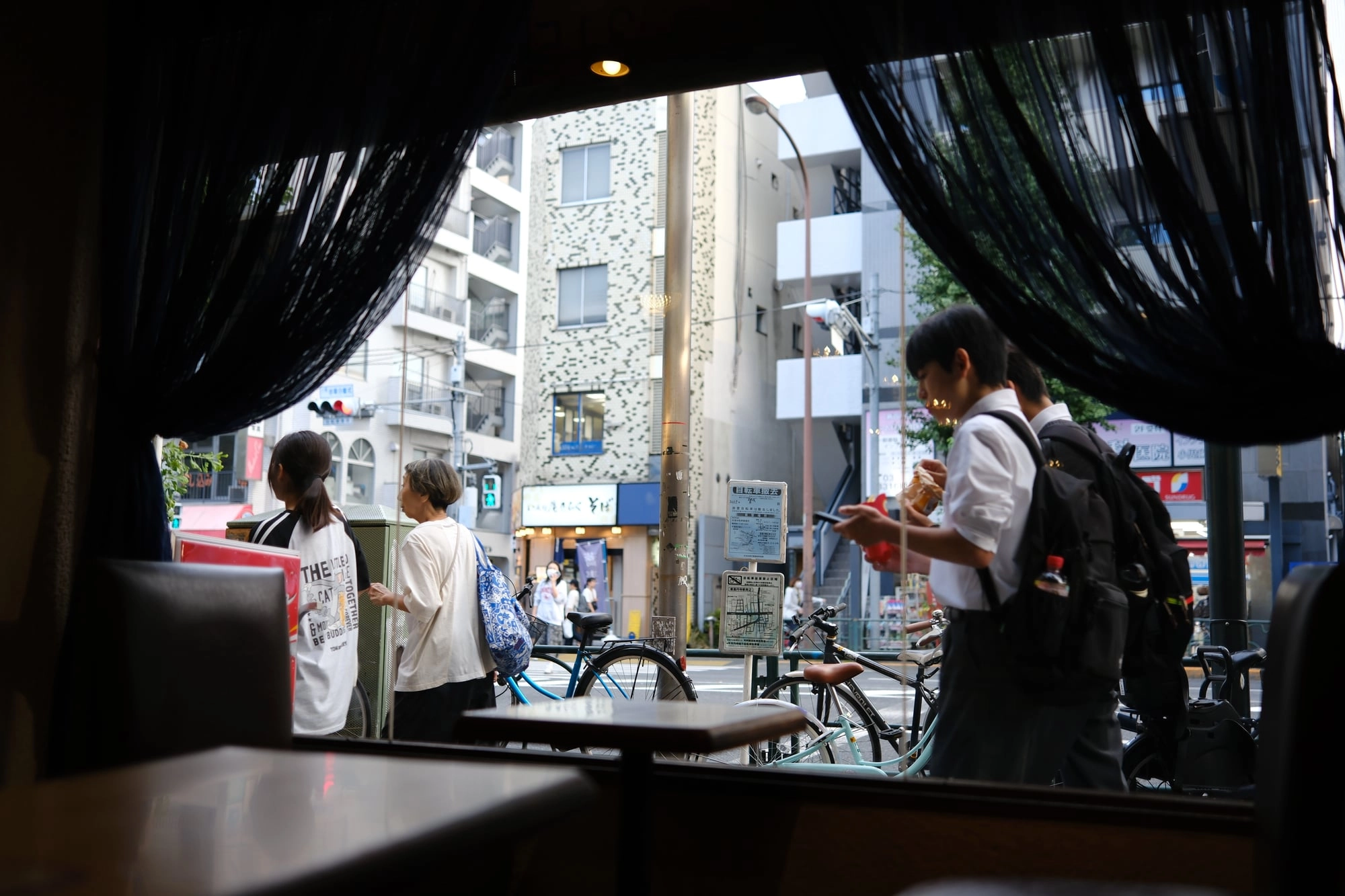 Framed by dark curtains, schoolboys and cyclists pause on a busy street outside a café window.