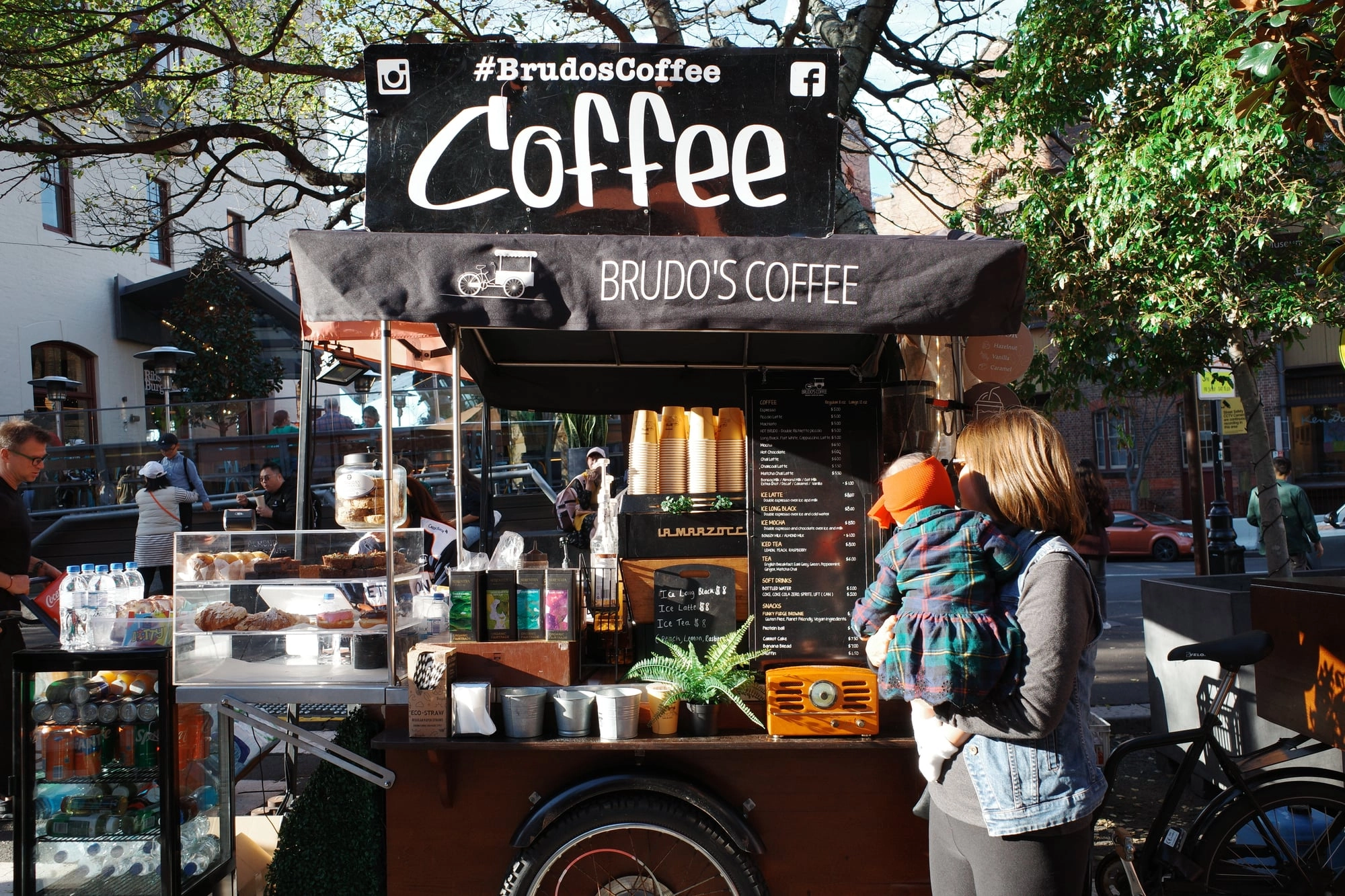 A woman holds a child in an orange hat while ordering at a bustling outdoor coffee cart.