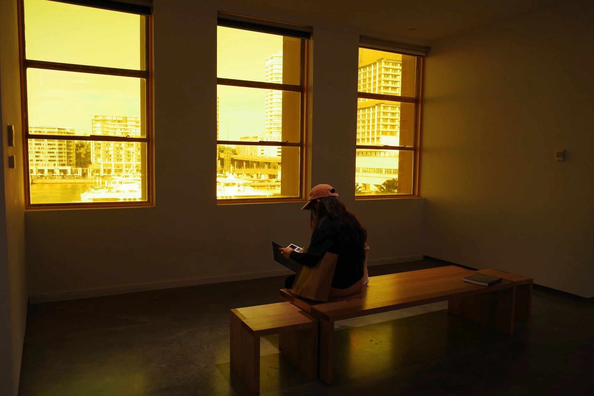 A seated woman looks out floor-to-ceiling windows at a harbor view bathed in golden light.