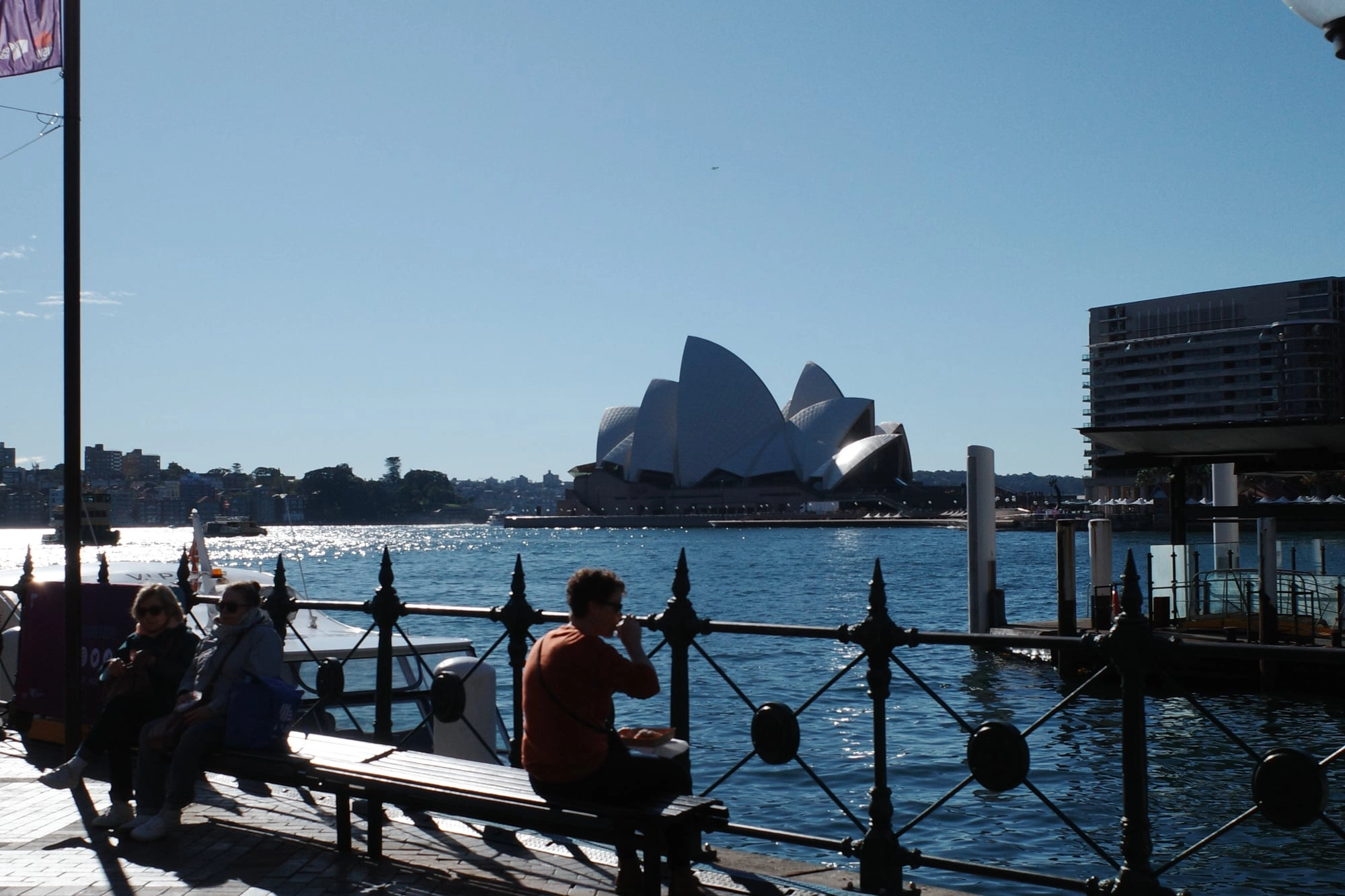 People relax on a waterfront bench across the harbor from the Sydney Opera House's white sails.