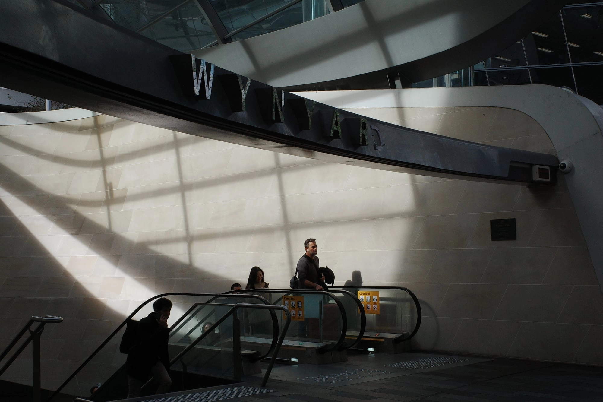 Commuters ride an escalator beneath a large station sign, lit by dramatic diagonal sunbeams.