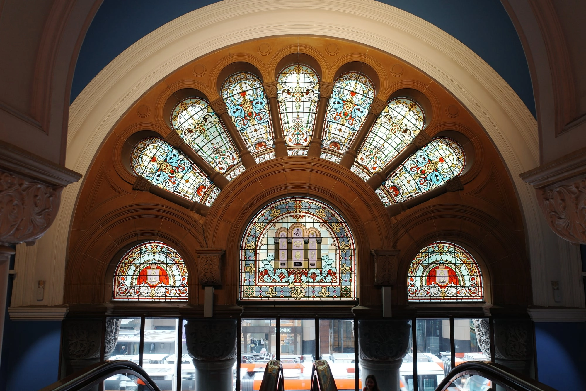 An ornate stained-glass fan window glows above an arched doorway in a grand historic hall.